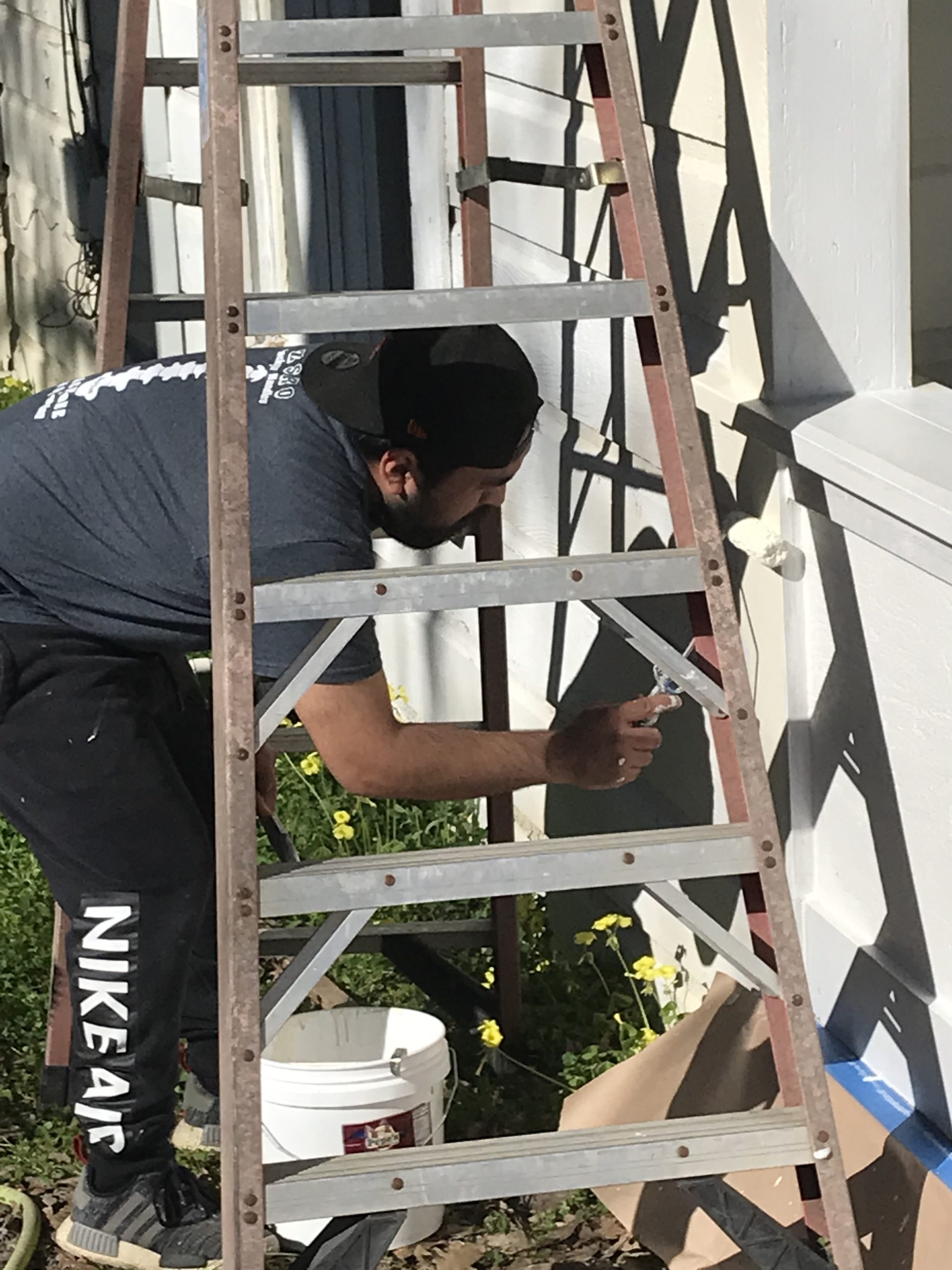 Latino (man) under a ladder painting the outside of a house.